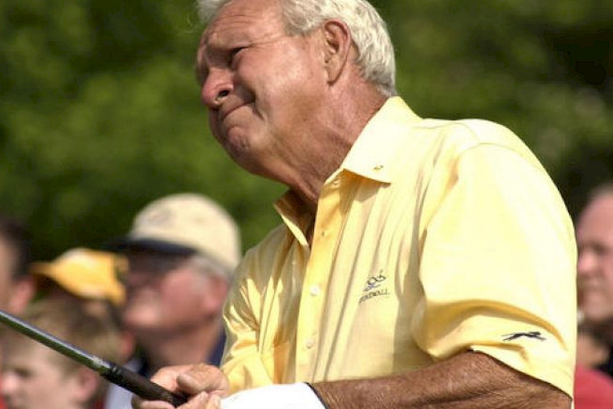 A person in a yellow shirt is holding a golf club, with spectators blurred in the background, suggesting a golf event setting.