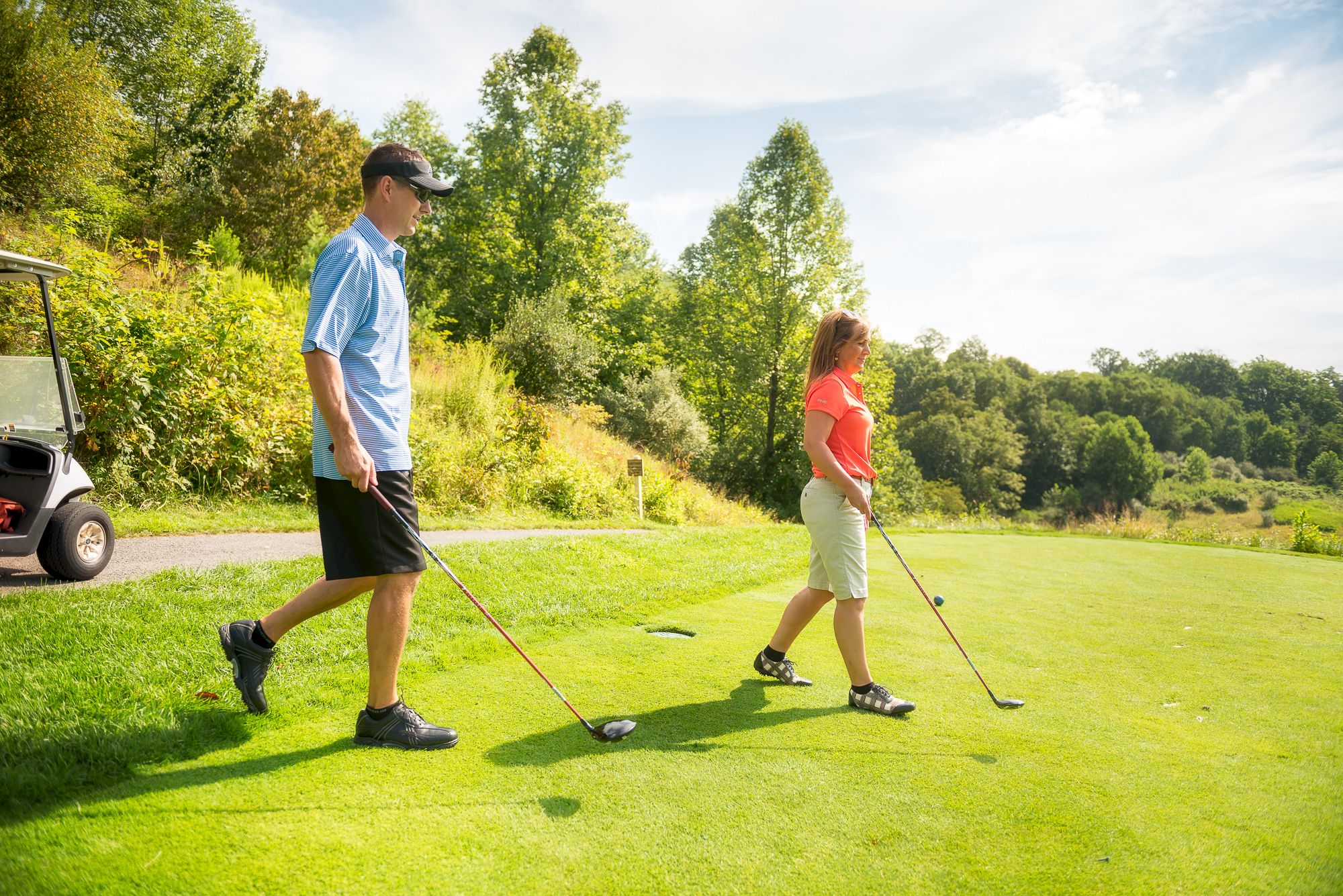 Two people are standing on a grassy area with golf clubs, near a golf cart, and surrounded by trees under a partly cloudy sky.