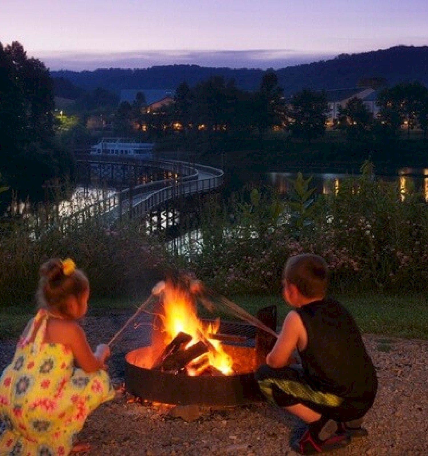 Two children are sitting near a campfire by a lake at dusk, with a scenic bridge and mountains in the background.