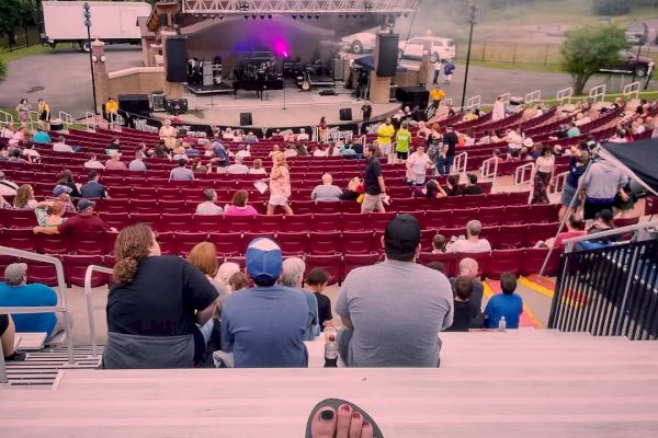 An outdoor concert venue with people seated in rows, a stage with sound equipment, and smoke, viewed from the perspective of someone with feet up.