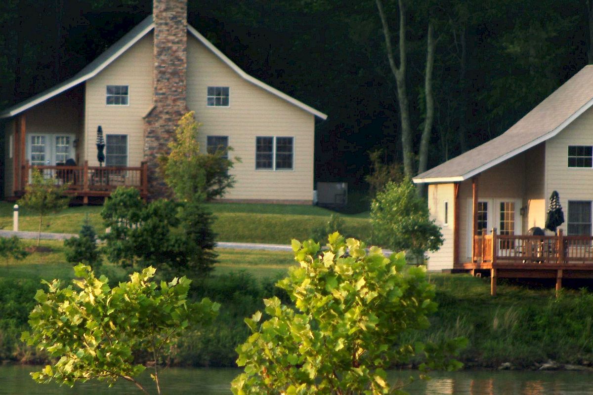 Two beige cottages with stone chimneys near a lake, surrounded by greenery and trees. Each cottage has a porch in the front.