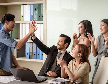 A group of five office workers are celebrating, with one person giving a high-five and others clapping and smiling around a table.