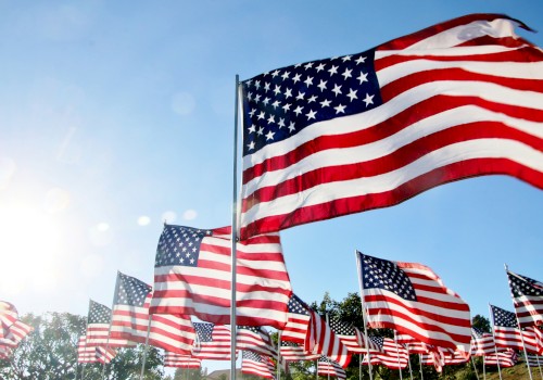 A group of American flags waving under a clear blue sky with the sun shining brightly in the corner of the image.