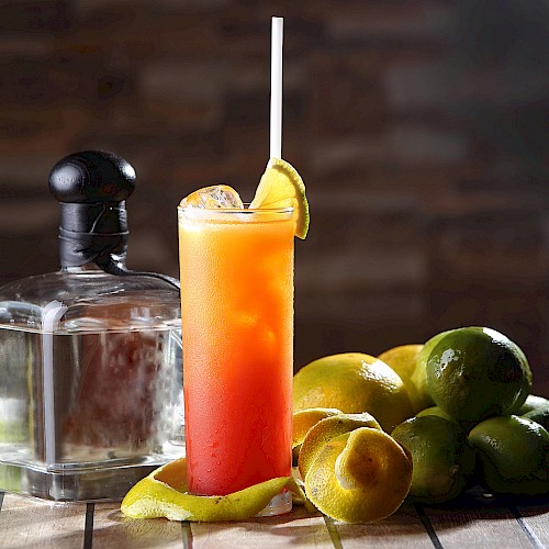 A cocktail with a straw and lime garnish, surrounded by limes and a liquor bottle on a wooden table.