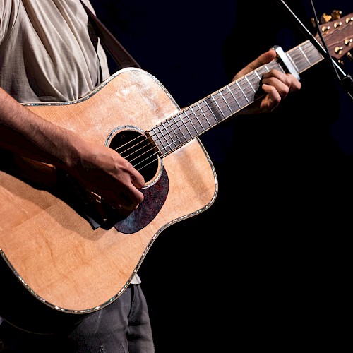 A person is playing an acoustic guitar on stage, holding a pick while wearing a casual shirt, with a microphone visible in the background.