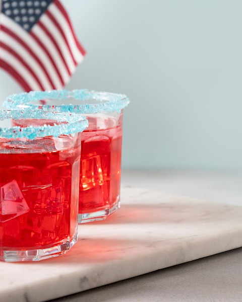 Two red drinks with sugared rims on a marble surface, accompanied by American flags in the background.