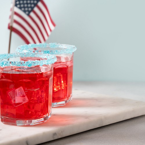 Two red drinks with sugared rims on a marble surface, accompanied by American flags in the background.