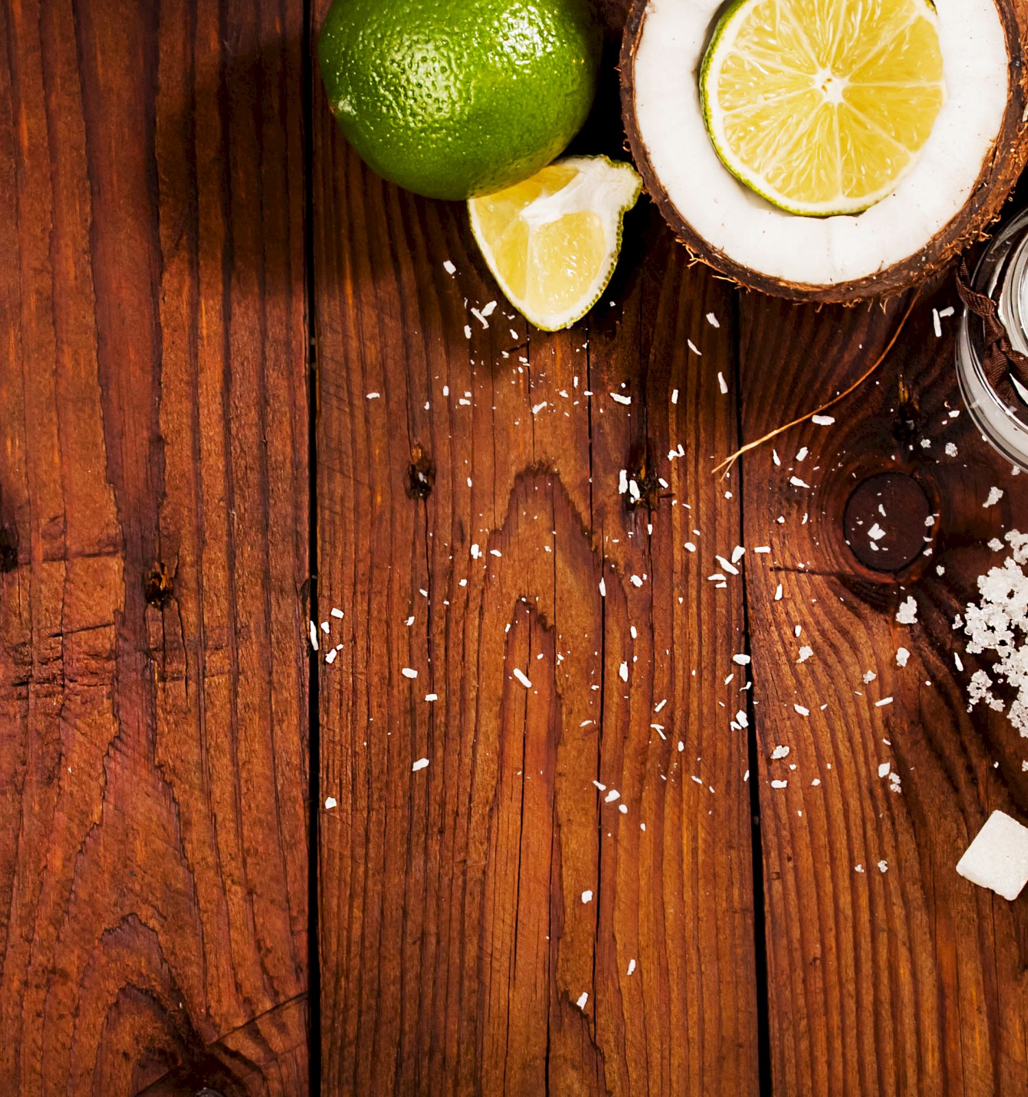 Coconut halves with limes, a jar of coconut milk, a spilled jar of sugar, sugar cubes, and lime slices on a wooden surface.