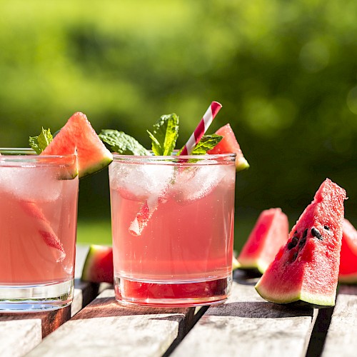 Two glasses of watermelon drinks with ice, garnished with watermelon slices and mint, placed on a wooden table outside.