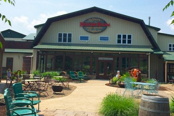 The image shows a rustic building with a sign on top, an outdoor seating area with tables and chairs, and greenery around.