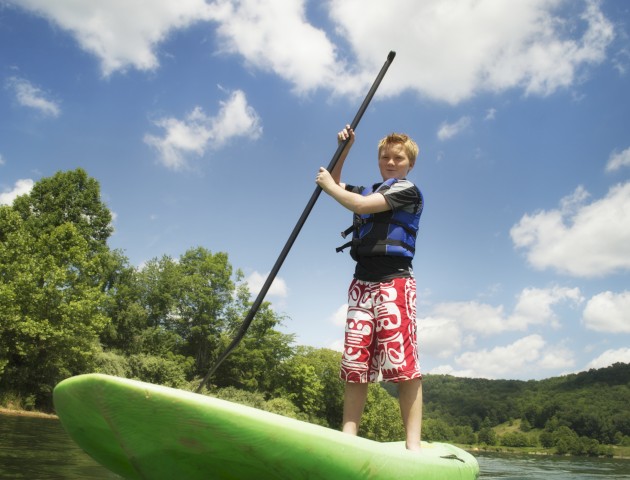 A person paddleboarding on a body of water, wearing a life jacket and red shorts, with trees and a blue sky with clouds in the background.