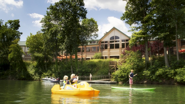 People enjoying a sunny day on the water with paddle boats, surrounded by greenery and a building in the background.