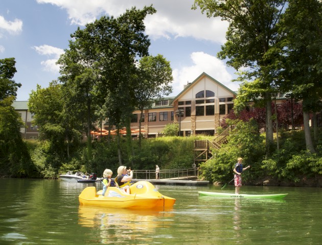 People enjoying water activities in a yellow paddle boat and green kayak on a lake, with a building and trees in the background.