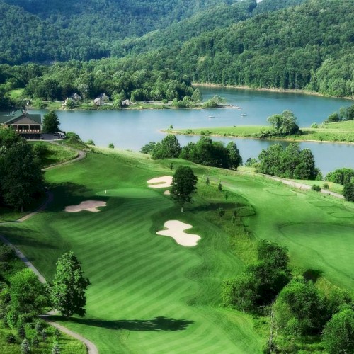 A scenic golf course with lush green fairways, sand traps, and a backdrop of a lake surrounded by forested hills under a clear sky.