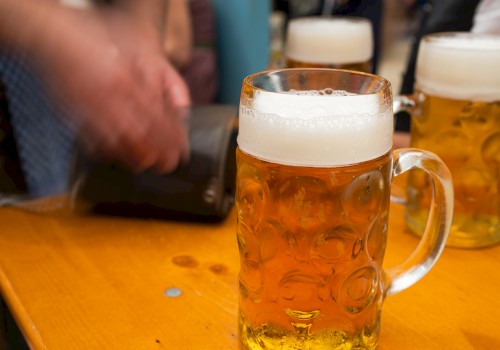 A table with large mugs of beer, one prominently in front. A person's blurred hand is near a wallet in the background.