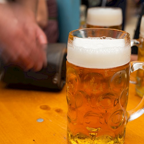 A table with large mugs of beer, one prominently in front. A person's blurred hand is near a wallet in the background.