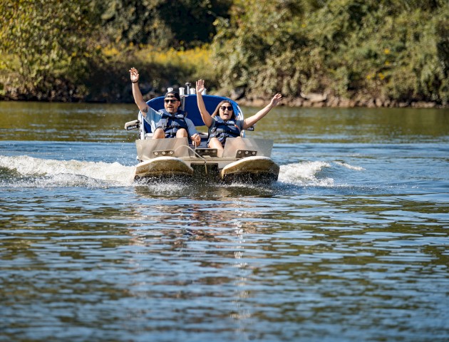 Two people wearing life jackets are enjoying a ride on a pedal boat in a scenic, sunny lake setting.