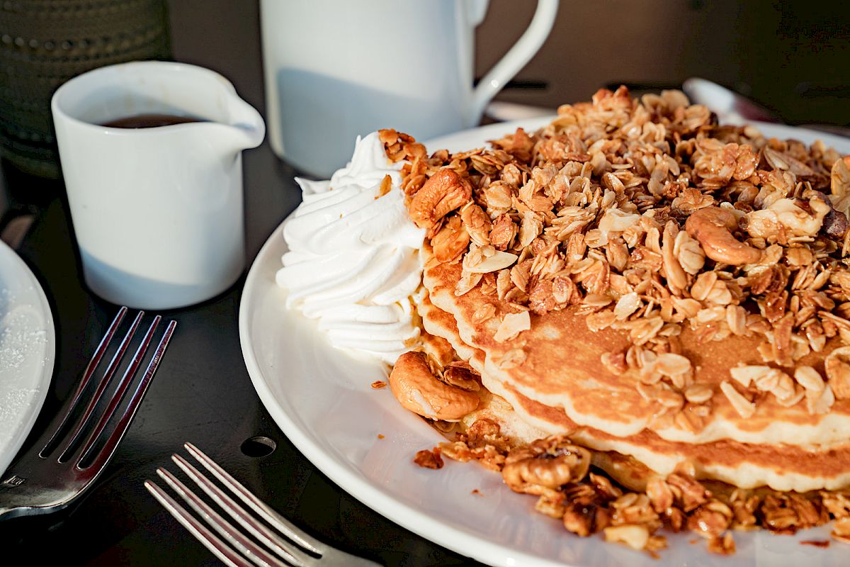 A stack of pancakes topped with granola and whipped cream, served with a jug of syrup on the side, alongside utensils.