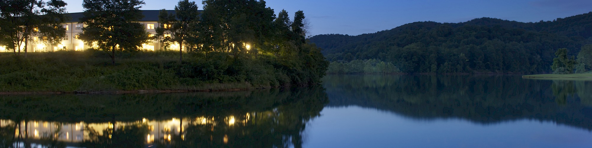 A serene lake at dusk with a building and trees reflected in the water, under a clear, deep blue sky.