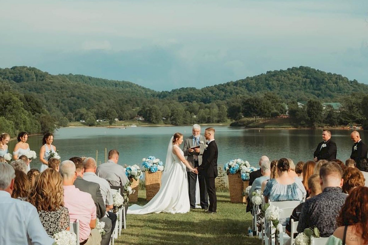A wedding ceremony by a lake with a couple exchanging vows. Bridesmaids and groomsmen stand on either side, guests seated watching the scene.