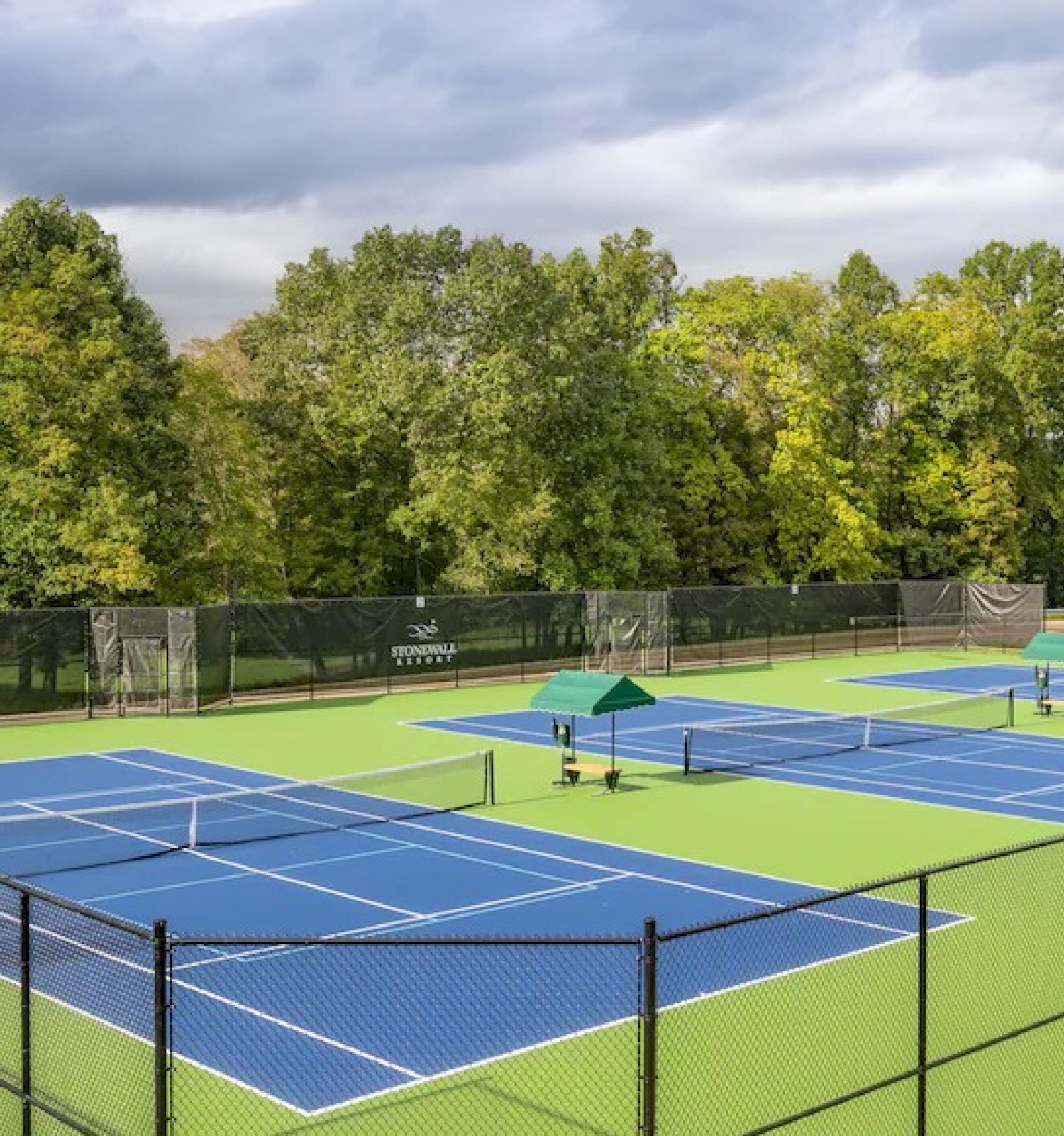 The image shows outdoor tennis courts surrounded by a fence and trees, with chairs and umbrellas beside the courts.
