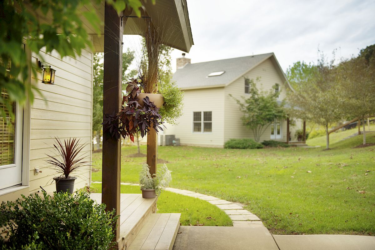 The image shows a peaceful suburban scene with a beige house, a porch with potted plants, a walkway, and a grassy yard with trees.