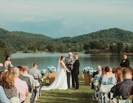 A wedding ceremony takes place outdoors by a lake, with guests seated and a scenic backdrop of hills and water, creating a serene setting.