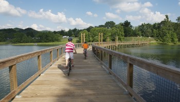 Two people bike across a wooden bridge over a lake, surrounded by greenery and under a partly cloudy sky.