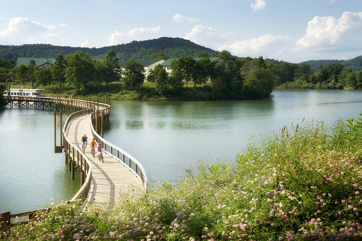 A curved boardwalk extends over a calm lake towards buildings, with people walking along it and green hills in the background under a blue sky.