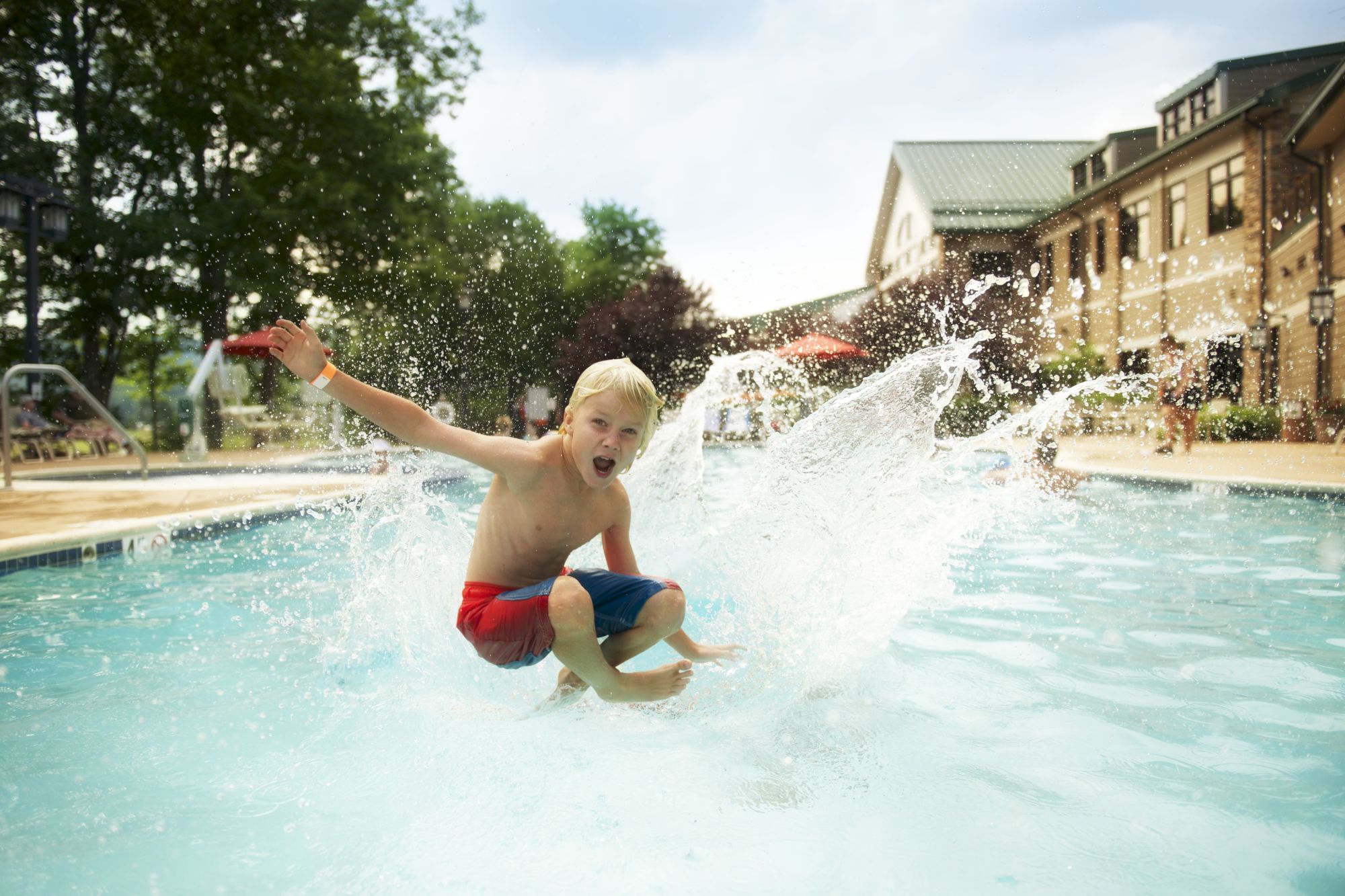 A child in red and blue shorts jumps into a pool, making a big splash, surrounded by a sunny outdoor setting with a building nearby.