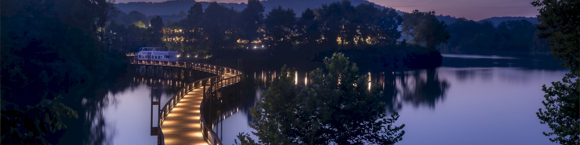 A wooden walkway over calm water, surrounded by trees and hills during twilight, with soft lights illuminating the path, ending the sentence.