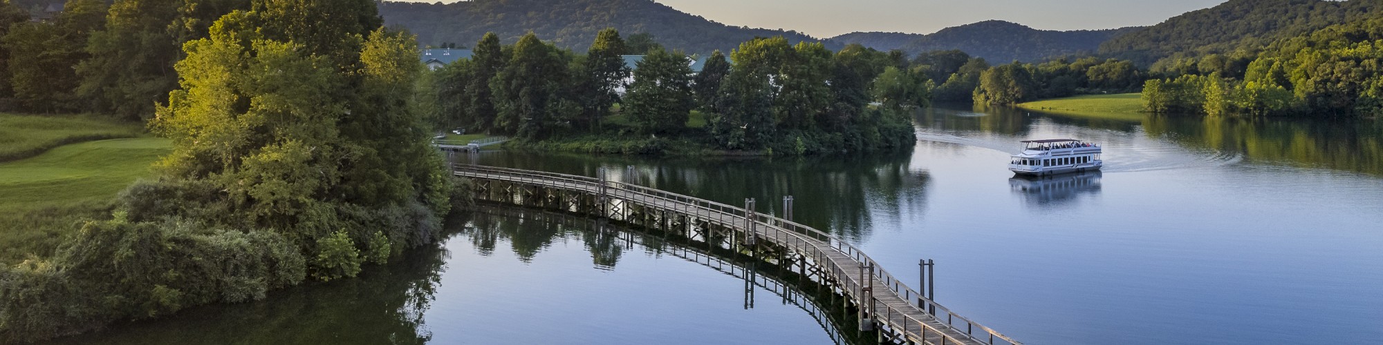 A serene lakeside view with a winding boardwalk, trees, hills, and a small boat on the tranquil water under a clear sky.