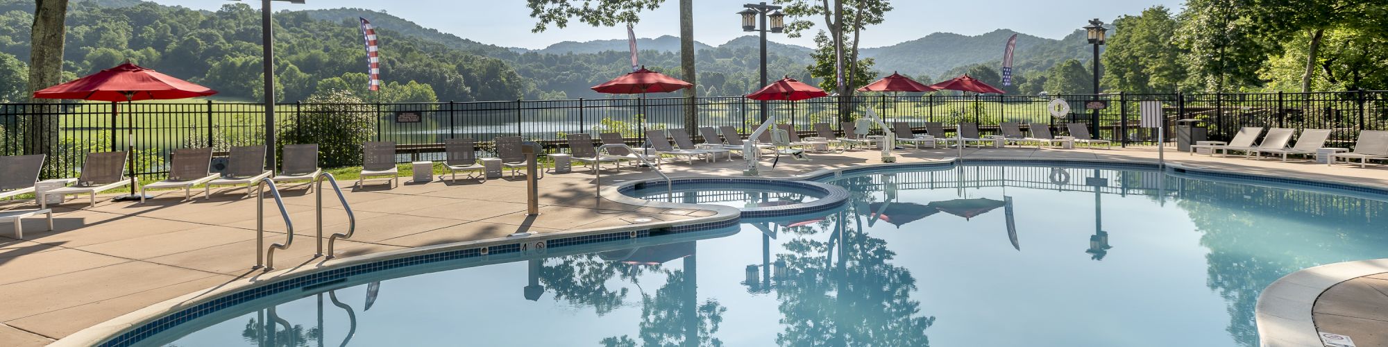 An outdoor swimming pool with clear water, surrounded by lounge chairs and red umbrellas. In the background are trees and mountains.