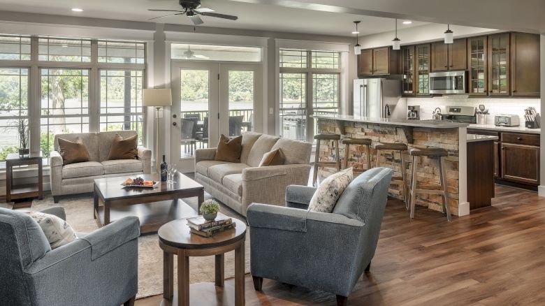 A modern living room and kitchen area with grey and beige furniture, wooden flooring, a ceiling fan, and large windows letting in ample light.