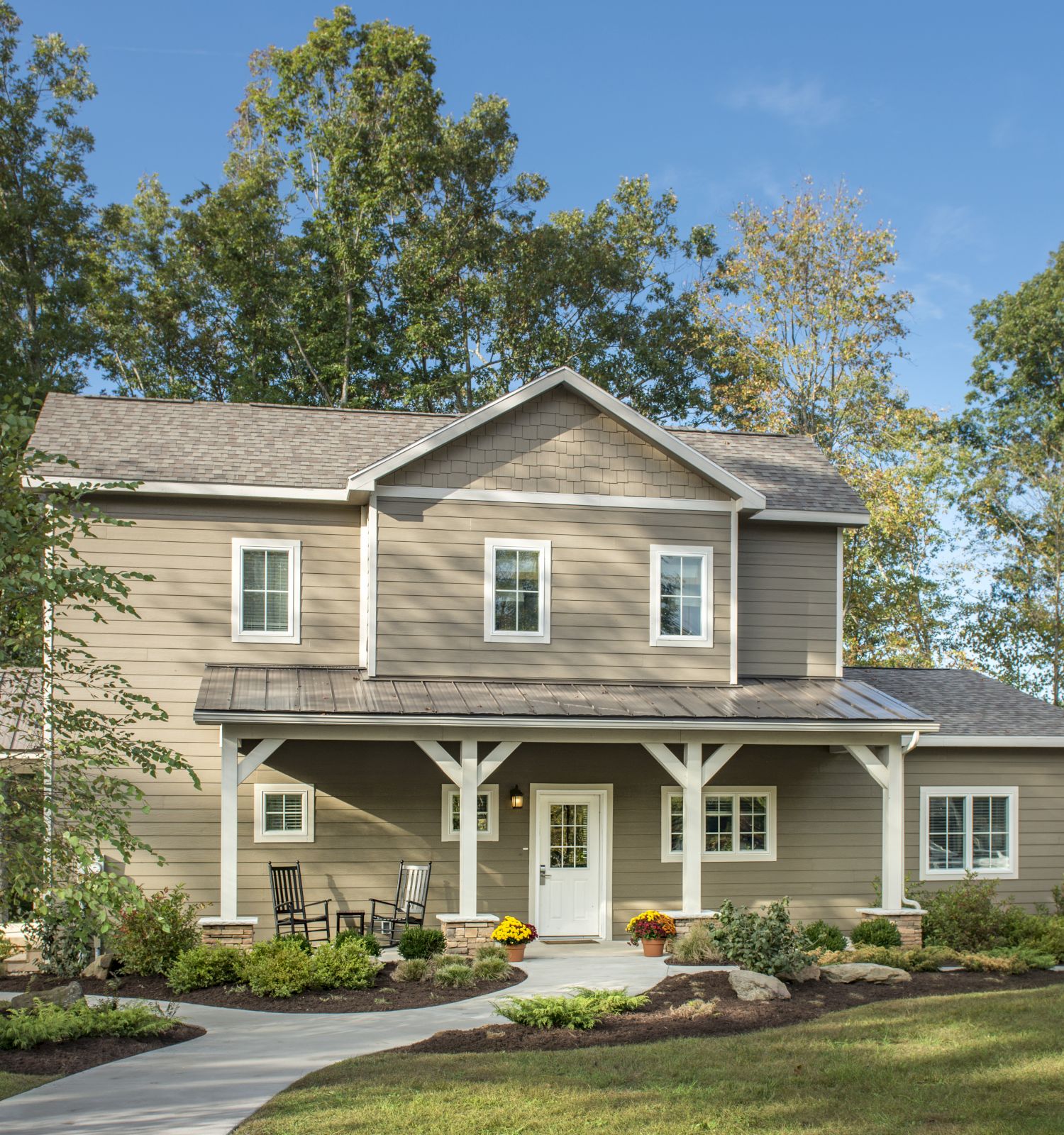 A two-story house with gray siding, white trim, and a covered porch surrounded by landscaped gardens and trees under a clear blue sky.