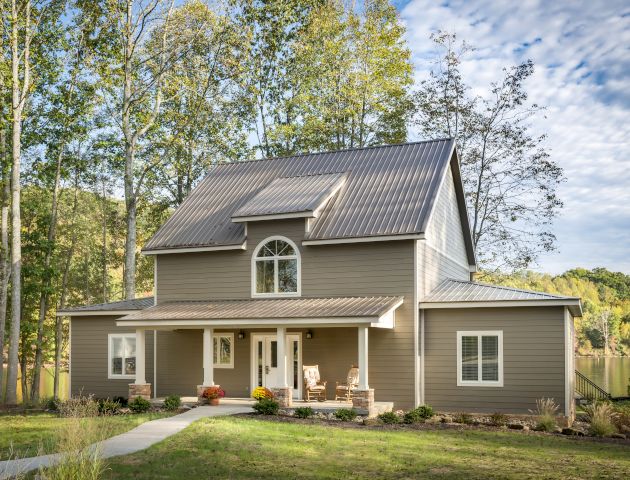 A two-story house with a metal roof, surrounded by trees and a lake in the background, features a porch with potted plants and outdoor seating.