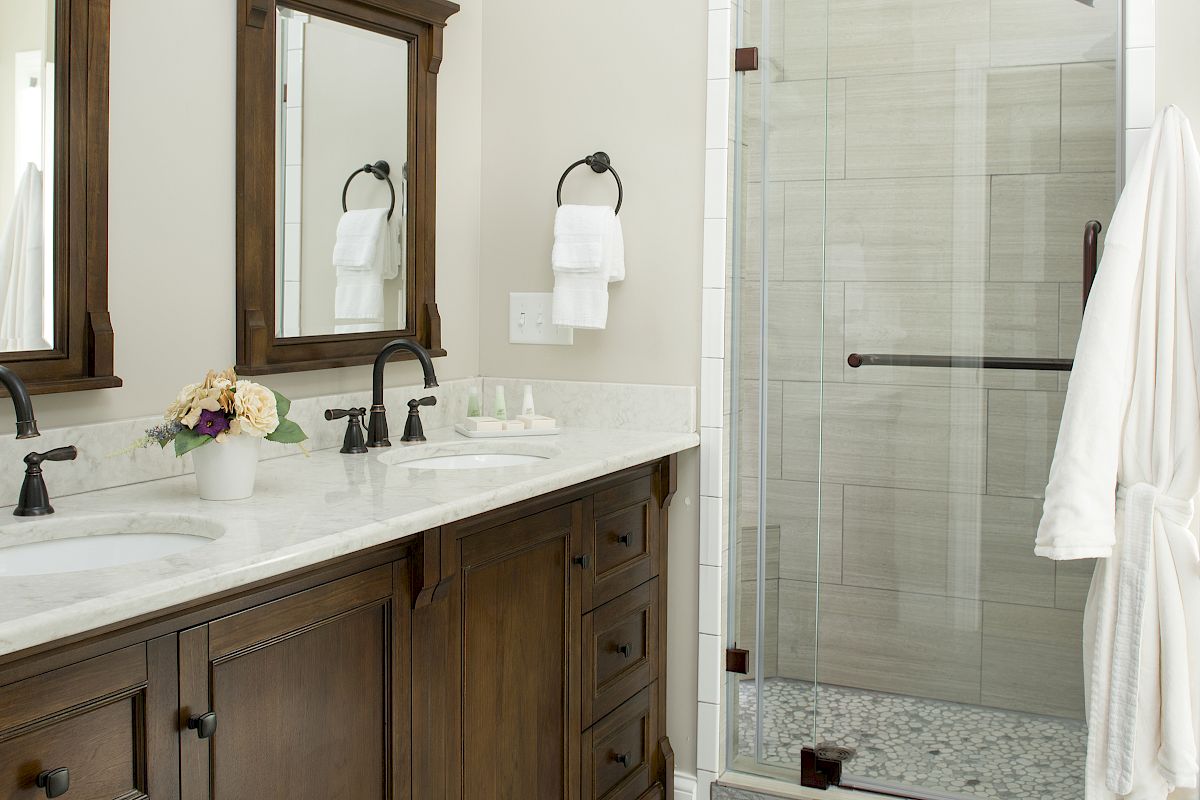 A modern bathroom with a wooden double vanity, marble countertop, wall-mounted lights, mirrors, and a glass-enclosed shower.