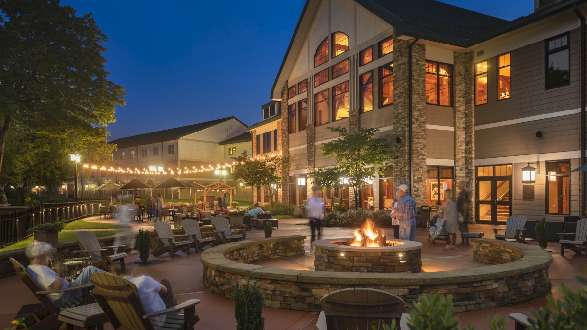 The image shows an evening outdoor scene at a resort with people gathered around a fire pit, surrounded by string lights, lounge chairs, and a large building.