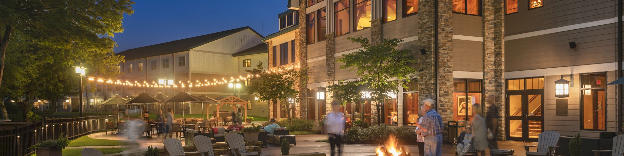 The image shows an outdoor seating area with a fire pit, surrounded by people and decorated with string lights, adjacent to a large building at night.