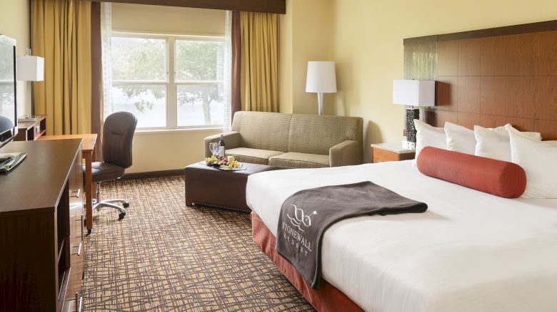 This image shows a well-lit hotel room with a bed, sofa, work desk, and TV, featuring large windows and warm-toned decor.