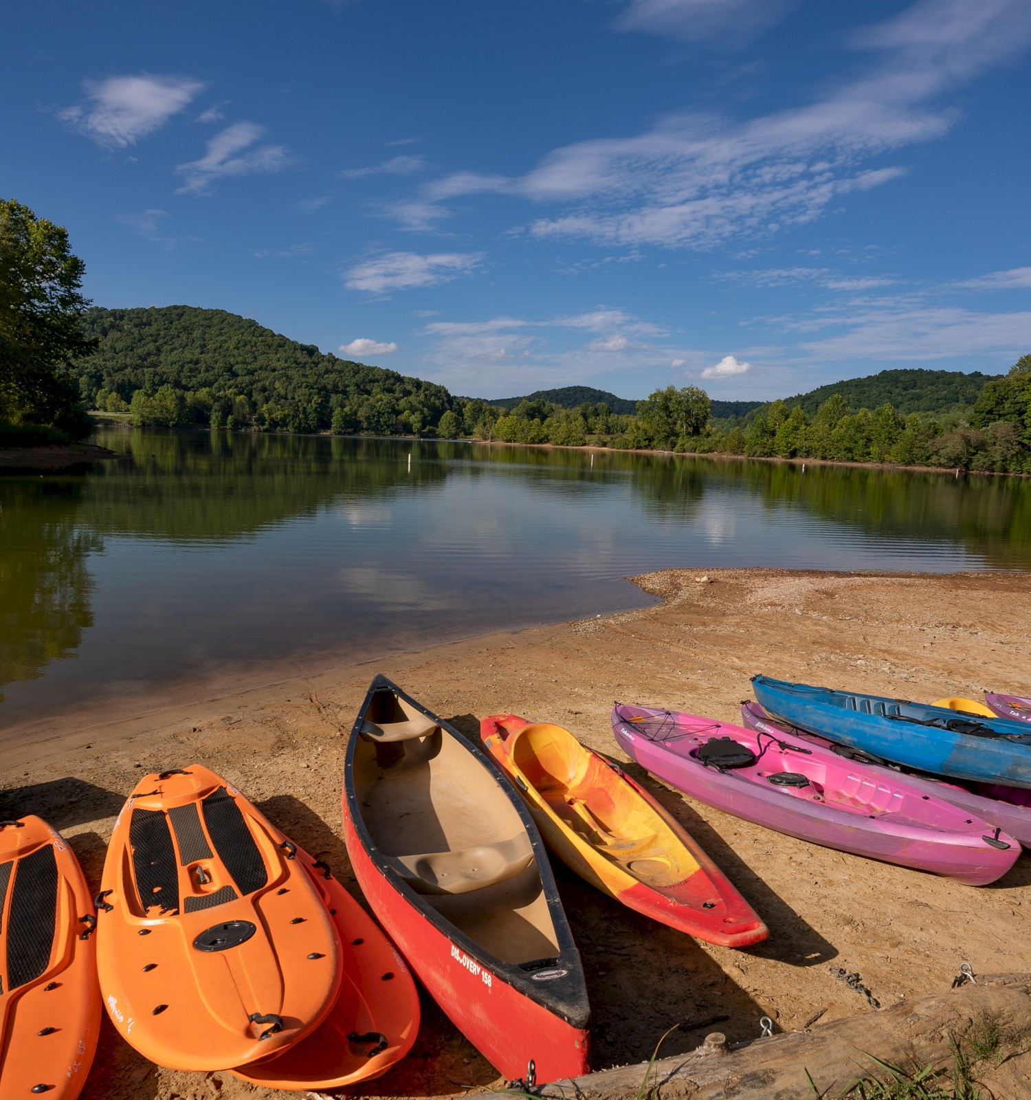 A picturesque lake scene with colorful kayaks and paddleboards on a sandy shore, surrounded by lush green hills and a clear blue sky.
