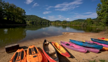 Kayaks and paddleboards are lined up on a sandy lakeshore, surrounded by green hills and under a blue sky with scattered clouds.