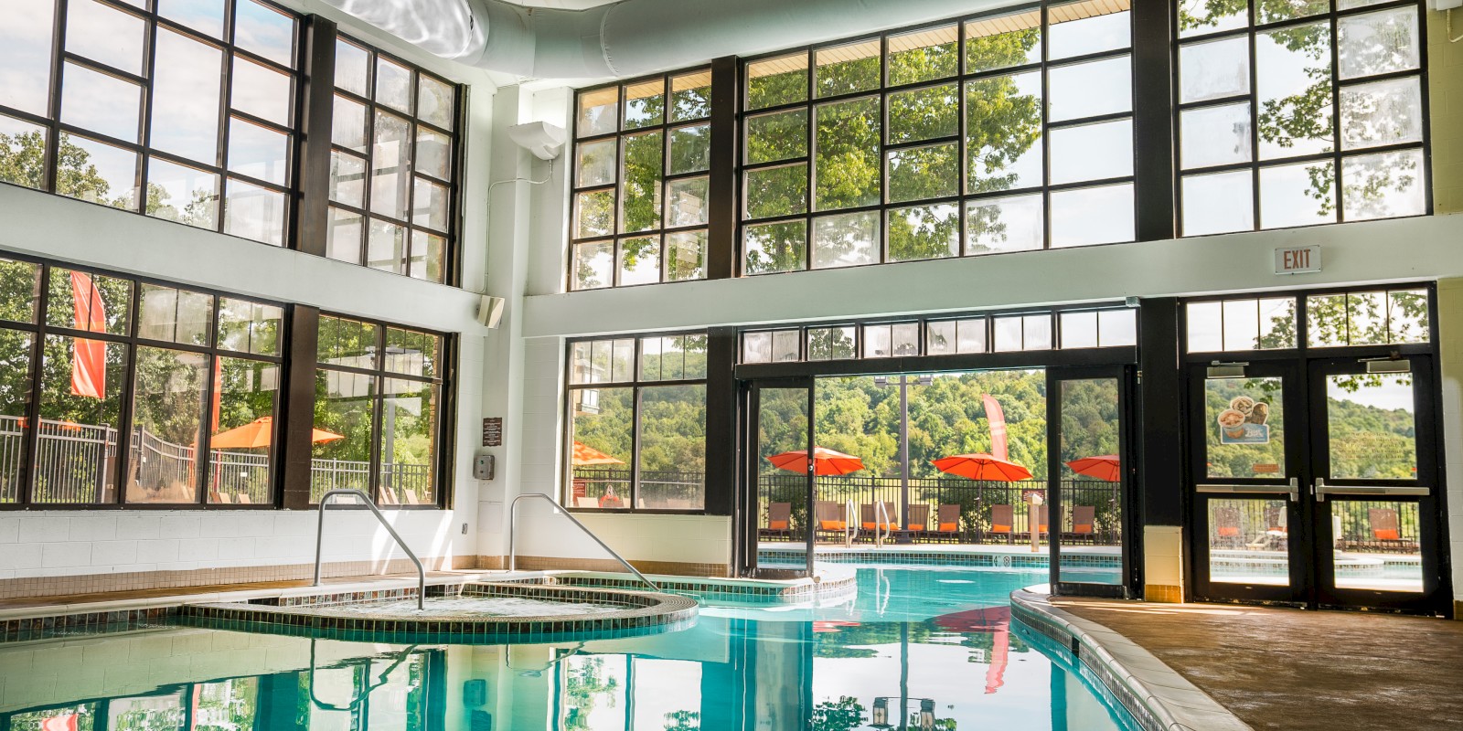 The image shows an indoor swimming pool with large windows, offering views of an outdoor area with umbrellas and greenery surrounding it.