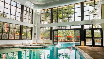 The image shows a brightly lit indoor swimming pool area with large windows and doors leading outside.