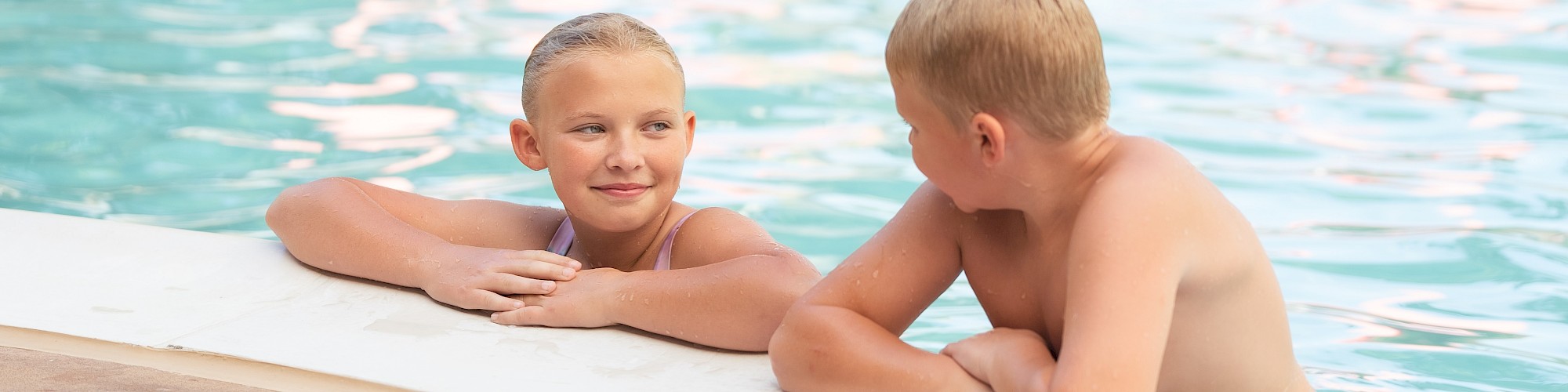 Two children are in a swimming pool, resting their arms on the pool's edge while having a conversation.
